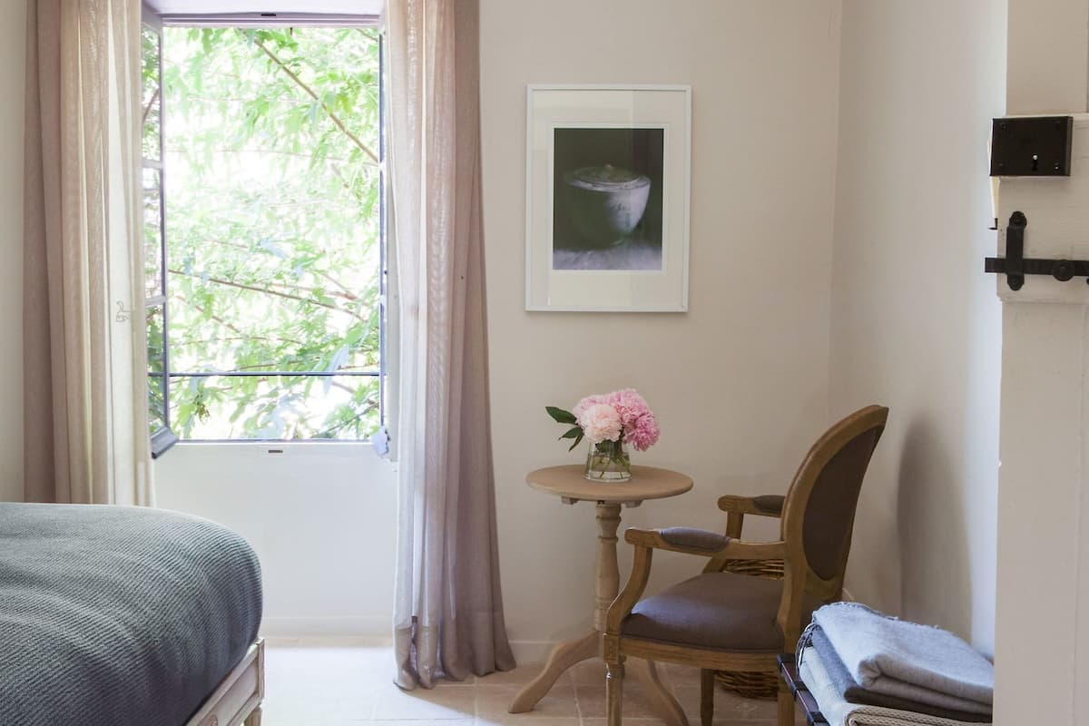 Une pièce comprenant un lit, une chaise, une petite table avec un vase de fleurs, un tableau encadré au mur et une fenêtre avec des rideaux.