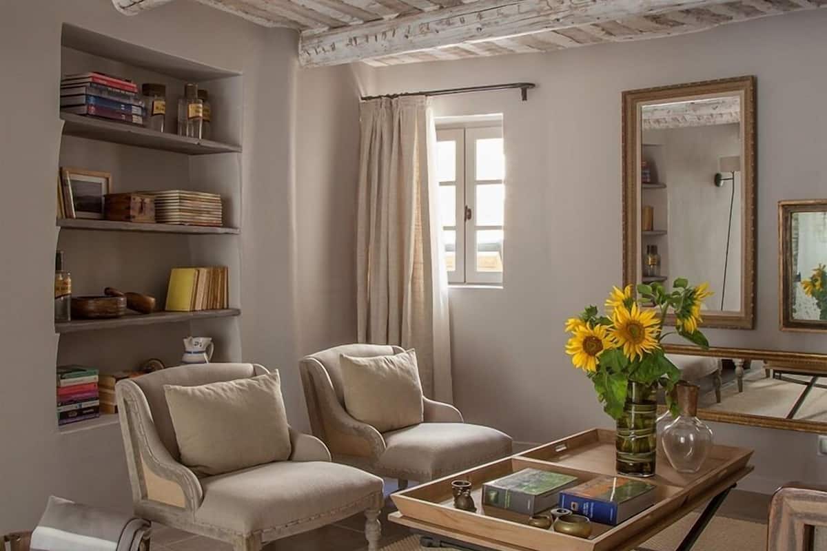 Un salon comprenant deux fauteuils, une table basse avec des livres et un vase de tournesols, une bibliothèque remplie de livres et d’objets décoratifs, et un grand miroir accroché au mur.