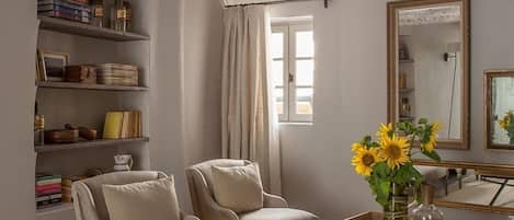 Un salon comprenant deux fauteuils, une table basse avec des livres et un vase de tournesols, une bibliothèque remplie de livres et d’objets décoratifs, et un grand miroir accroché au mur.