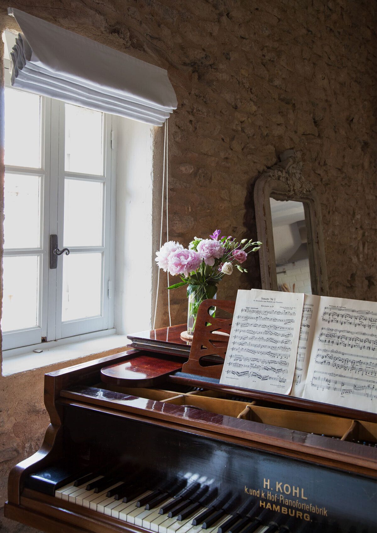 Un piano de cola con partituras, un florero con flores y un espejo en una habitación con paredes de piedra y una ventana.