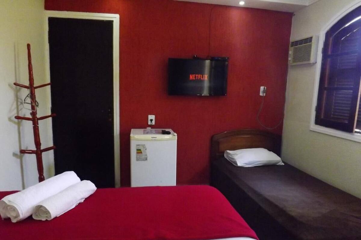 A hotel room with a bed, a small bedside table, a TV mounted on the wall, and a window with blinds.