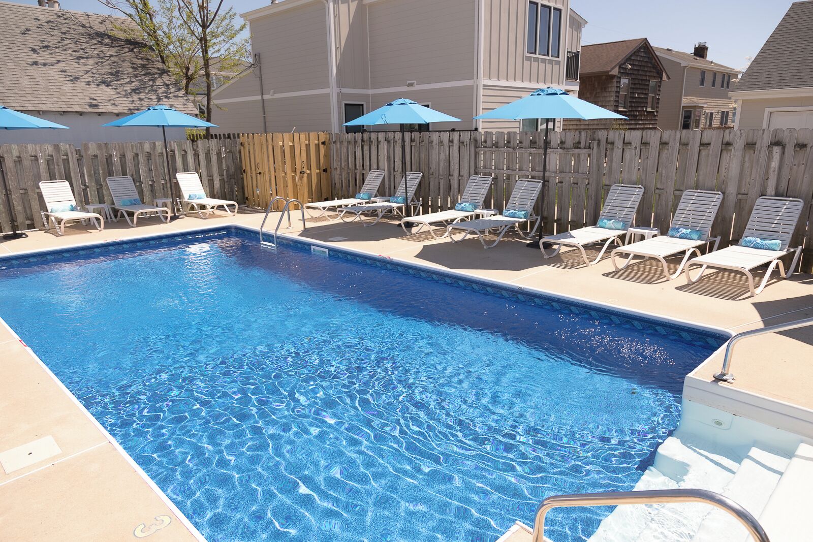 A rectangular swimming pool with a wooden deck and lounge chairs, surrounded by a wooden fence and umbrellas.