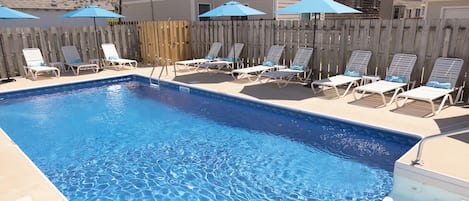 A rectangular swimming pool with a wooden deck and lounge chairs, surrounded by a wooden fence and umbrellas.
