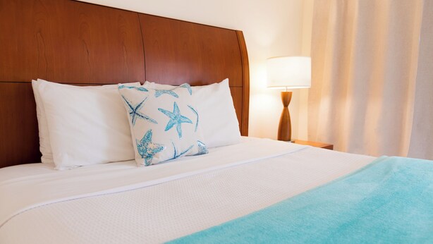 Ein Hotelzimmer mit einem Holzkopfteil, weißer Bettwäsche, einem Kissen mit Seestern-Muster, einer türkisblauen Tagesdecke und einer Nachttischlampe.