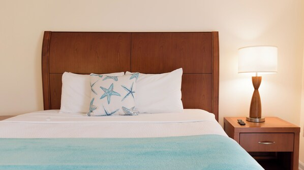 A hotel room with a wooden headboard, a bed with white bedding and a blue blanket, a nightstand with a lamp, and a drawer. - Broadway Court Motel (Point Pleasant Beach)