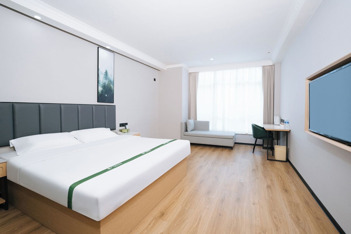 A hotel room with a bed, a desk, a chair, a window with curtains, and a large flat-screen TV.