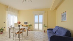 A dining area with a table and chairs, a blue sofa, and a television. - Residence La Nona (Cervia)