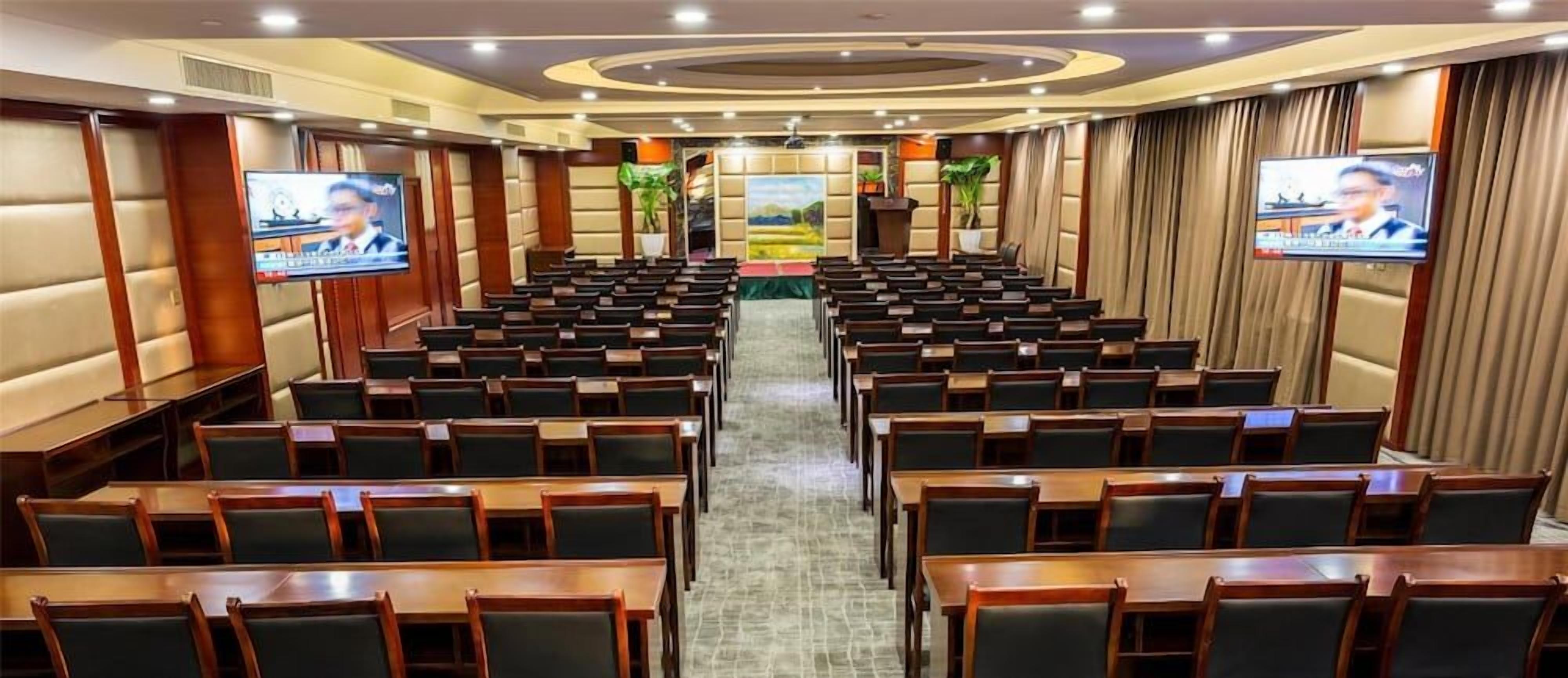 A conference room with rows of tables and chairs, a stage with a podium, and two screens on the walls.