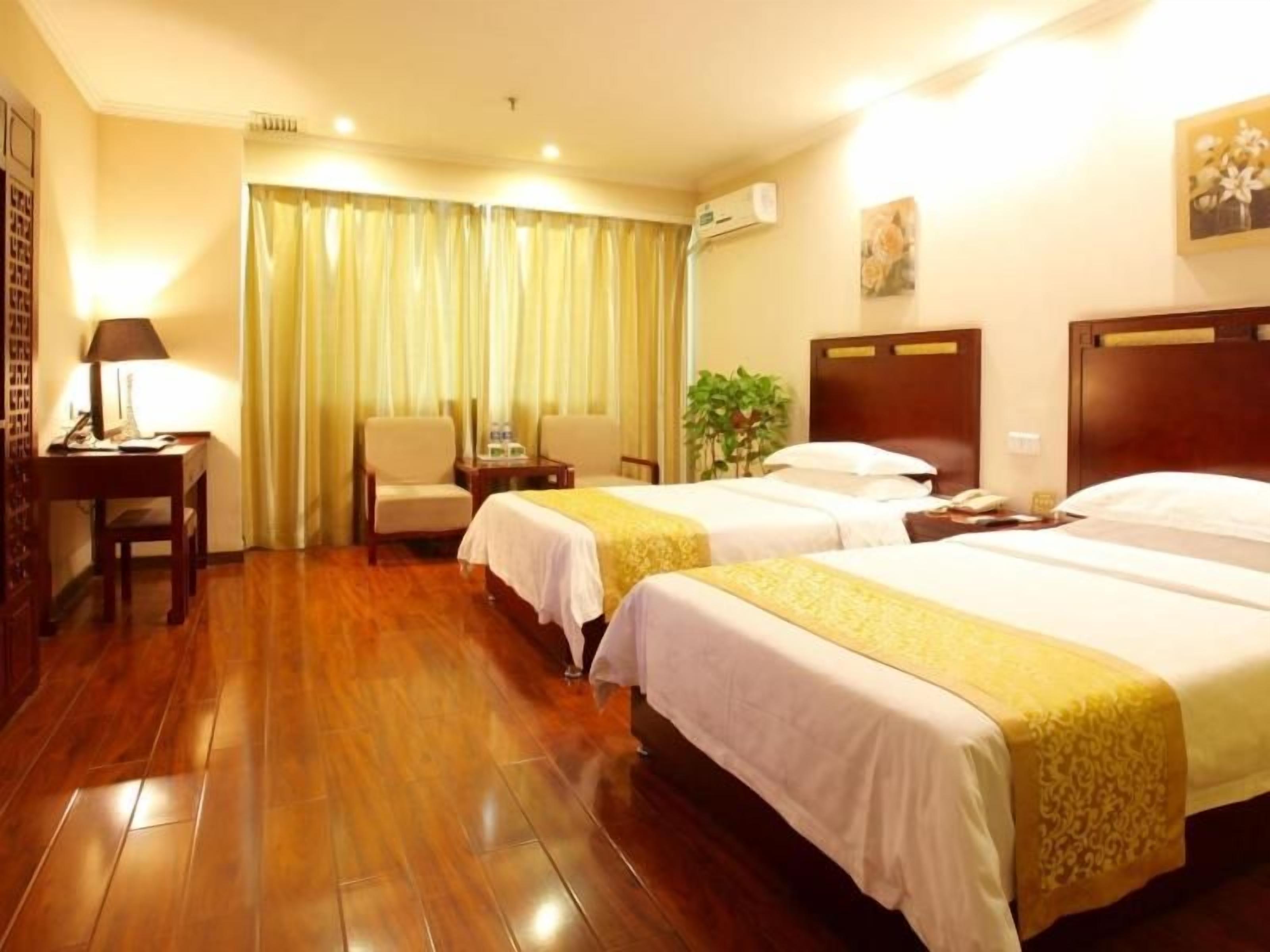 A hotel room with two beds, wooden flooring, a desk with a lamp, a chair, and a plant.