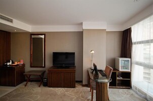 Habitación de hotel con televisión de pantalla plana, una mesita con una silla, minibar, espejo grande y ventana con cortinas.