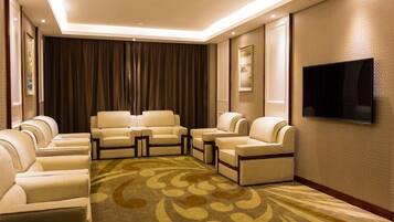 A hotel room with a seating area featuring sofas and armchairs, a flat-screen TV mounted on the wall, and a patterned carpet.