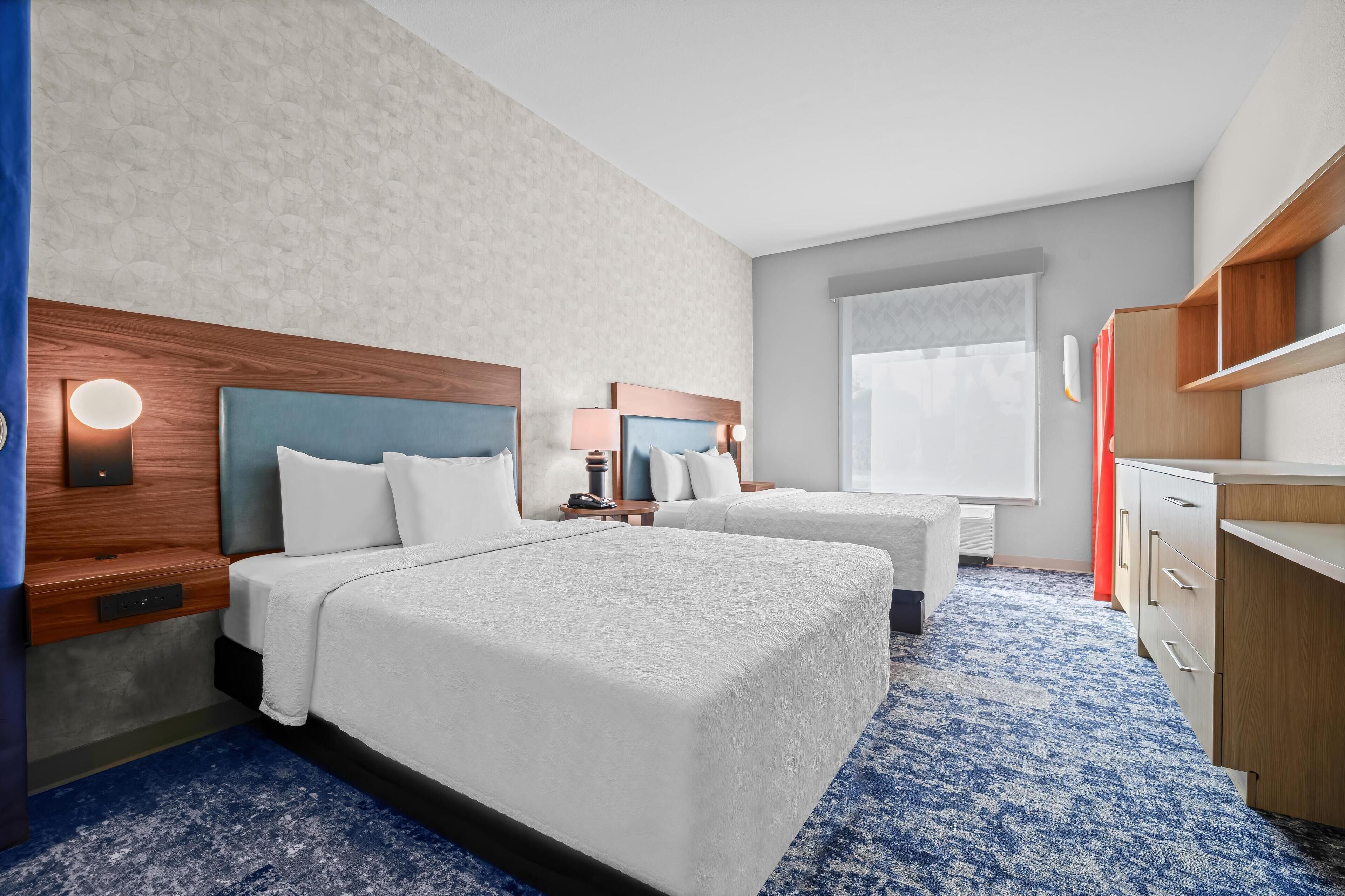 A hotel room with two beds, a wooden headboard, a nightstand with a lamp, a desk with drawers, and a large window.