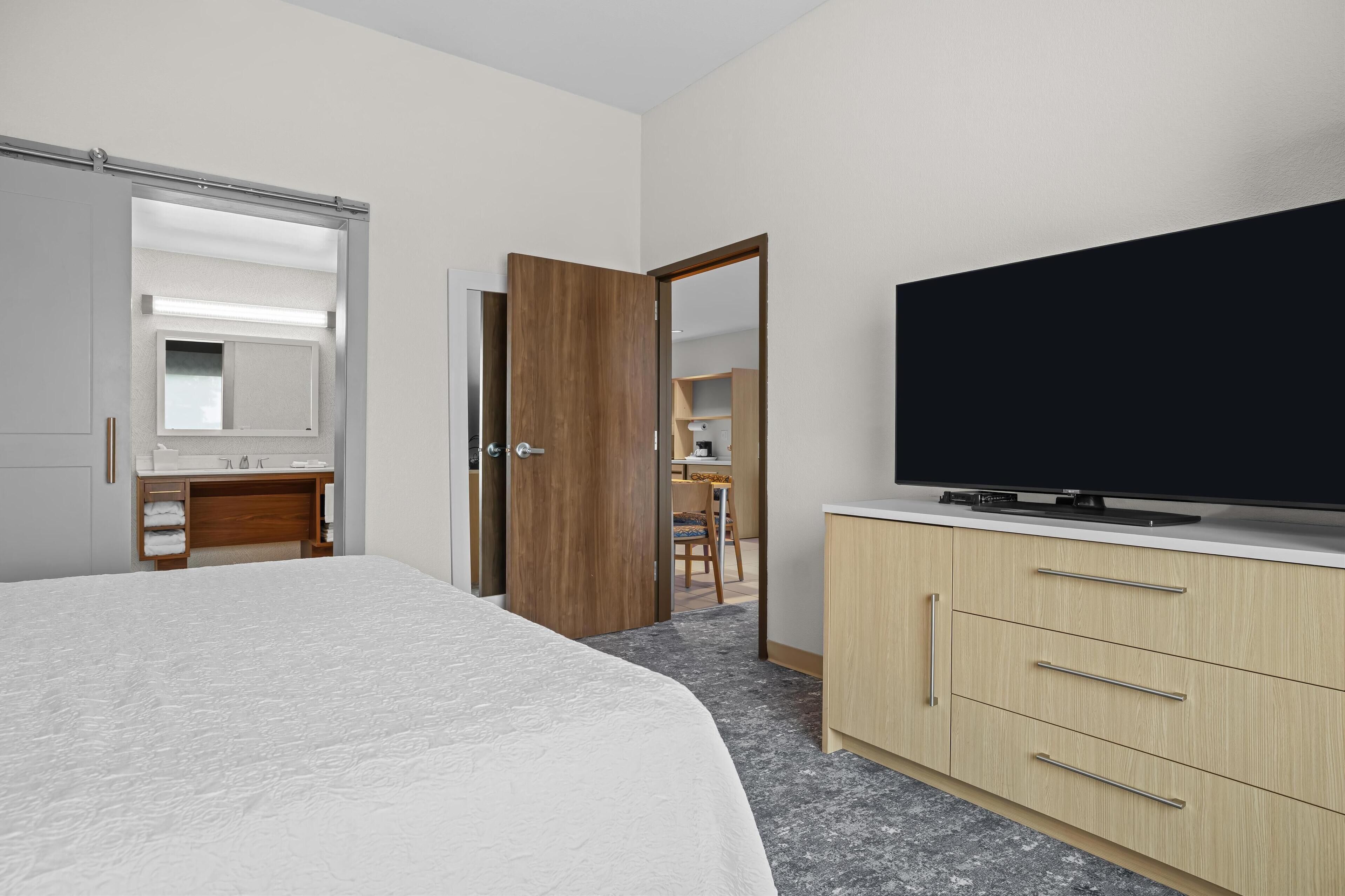 A hotel room with a bed, a TV, a wooden wardrobe, and a bathroom with a mirror and towel rack.