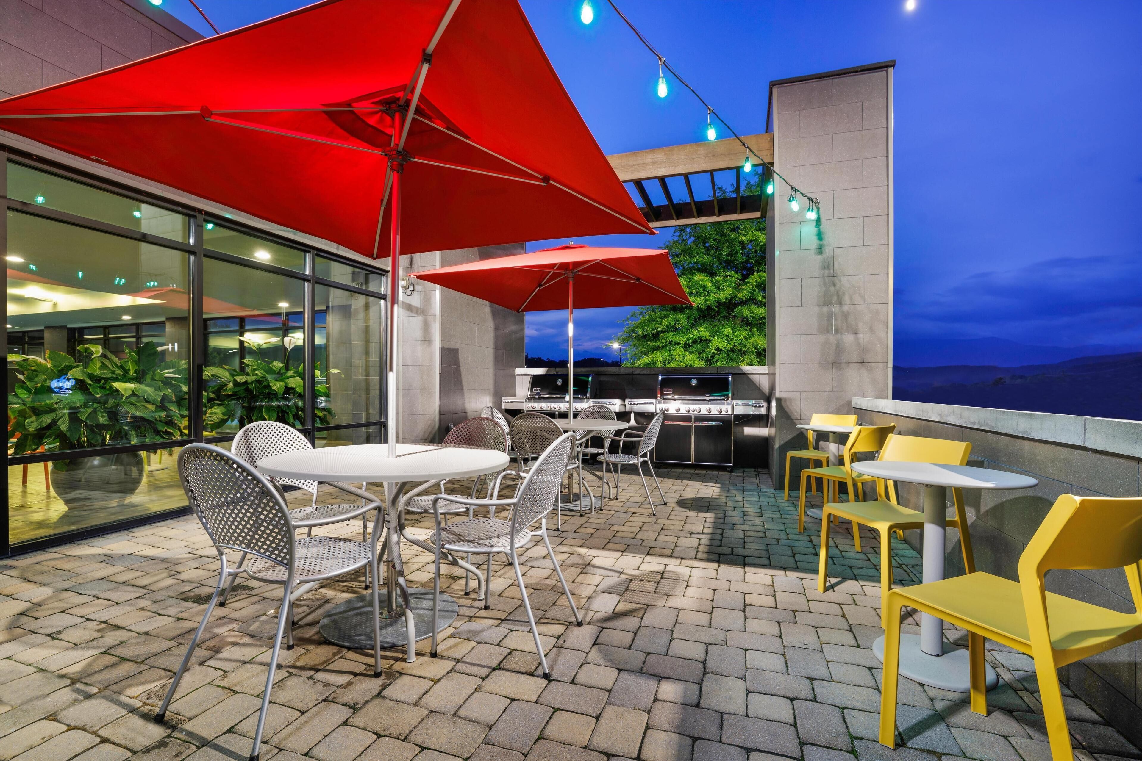 Eine Außenpatio mit Tischen, Stühlen, roten Sonnenschirmen und einem Grillbereich.