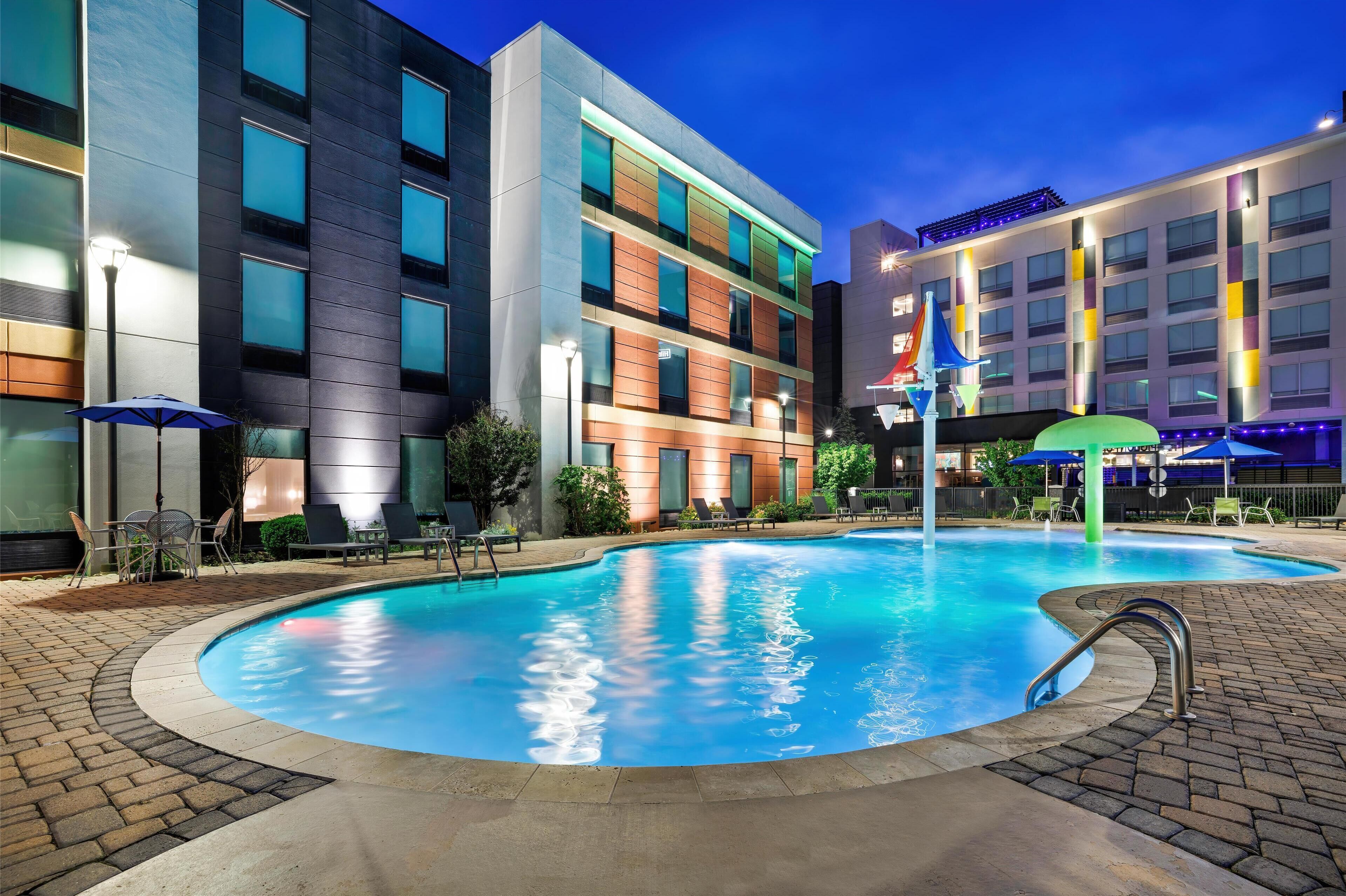 Et moderne hotel med swimmingpool, udendørs siddepladser og en velbelyst facade.