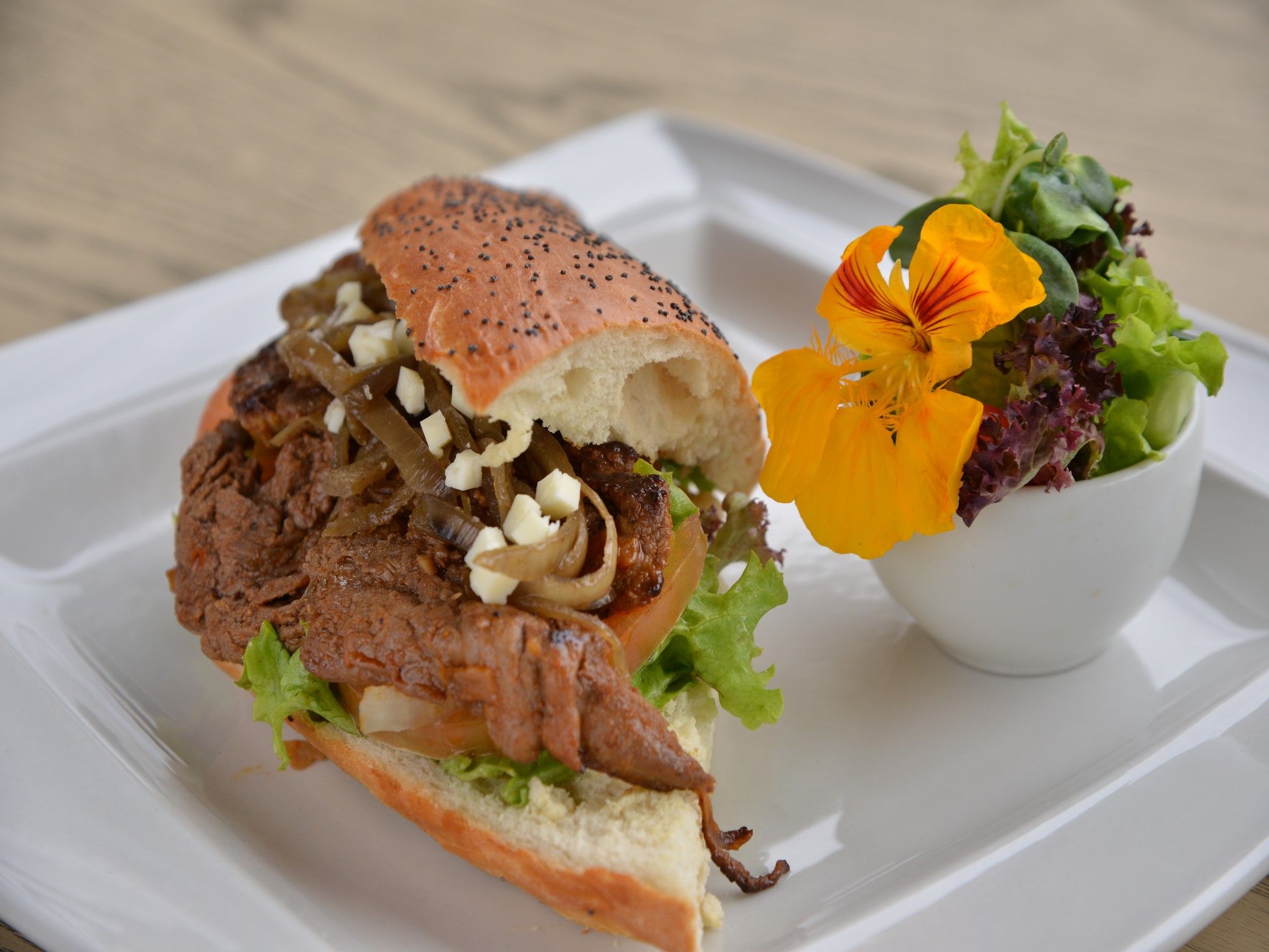 A sandwich with meat, onions, and cheese on a bun, accompanied by a side salad with a flower.