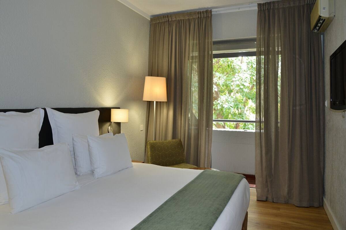 A hotel room with a large bed, a bedside lamp, a window with curtains, and a wall-mounted TV.