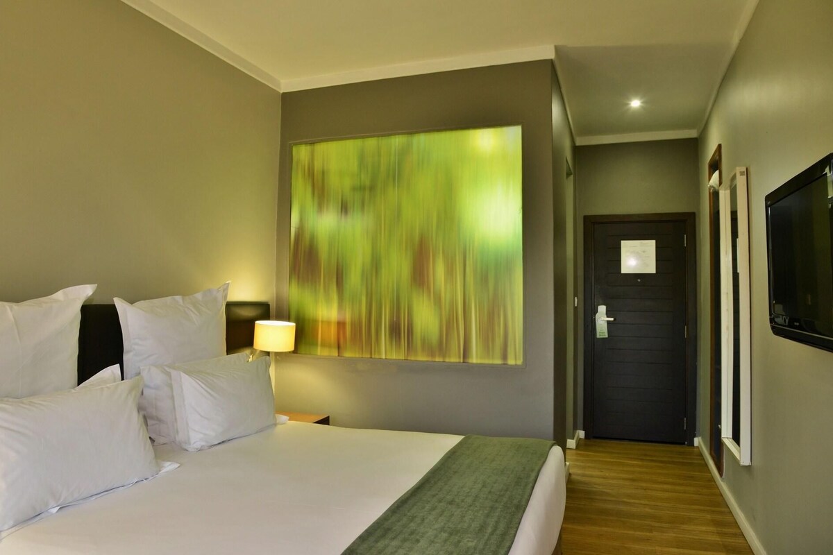 A hotel room with a large bed, a nightstand with a lamp, a green abstract painting on the wall, a door, and a TV mounted on the wall.