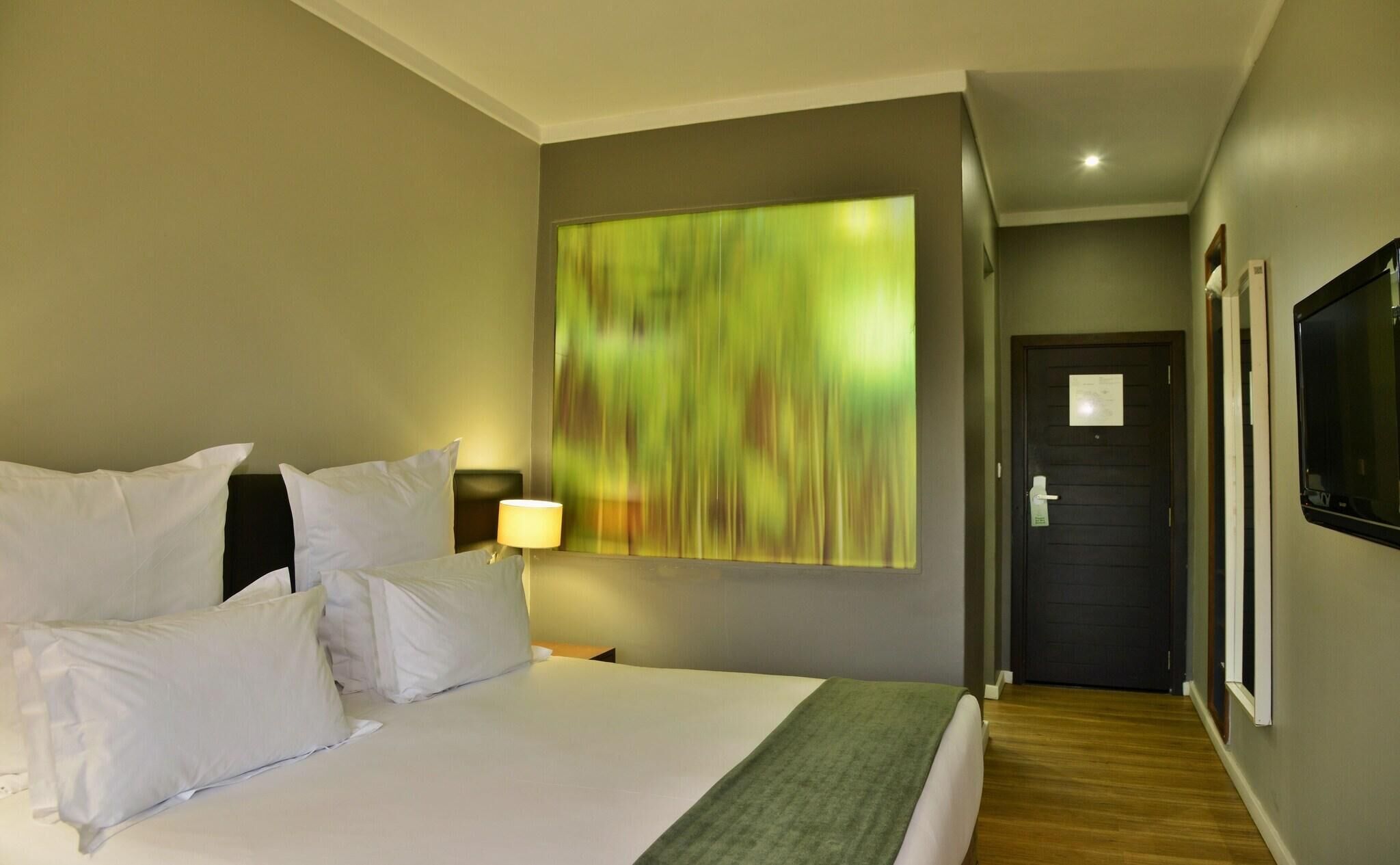 A hotel room with a large bed, a nightstand with a lamp, a green abstract painting on the wall, a door, and a television mounted on the wall.