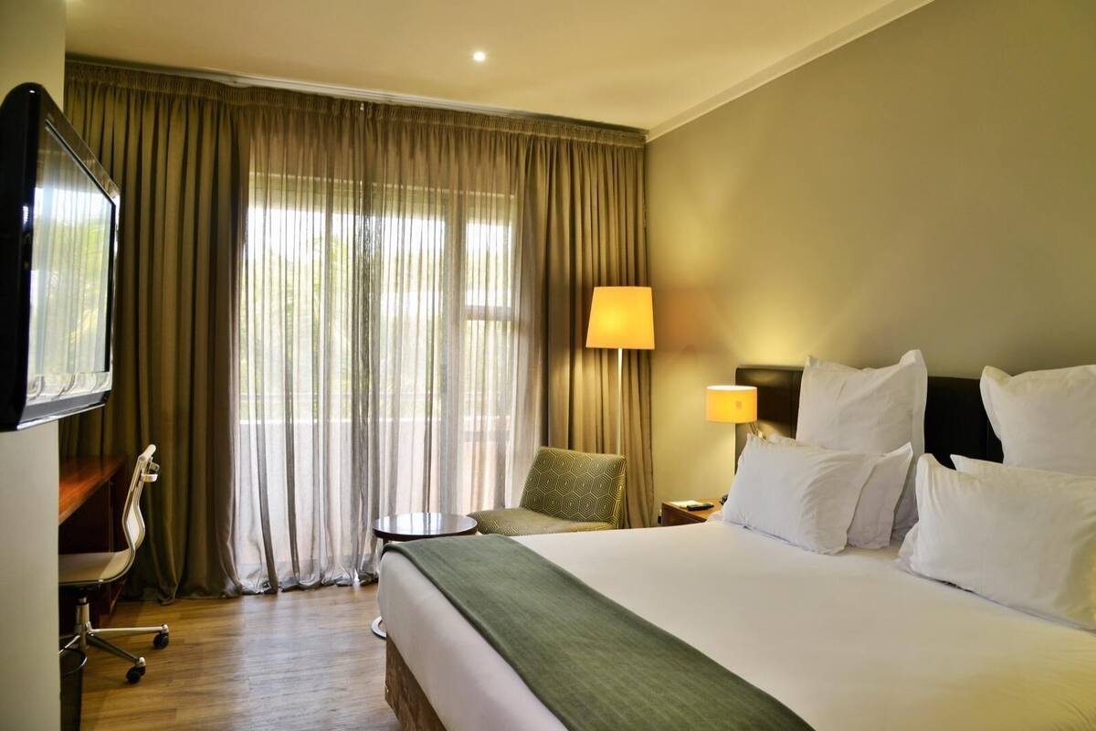 A hotel room with a large bed, two bedside lamps, a TV, a desk with a chair, and a window with curtains.
