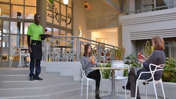 An outdoor seating area with tables and chairs, a waiter serving a drink, and a building with large windows and a sign in the background.