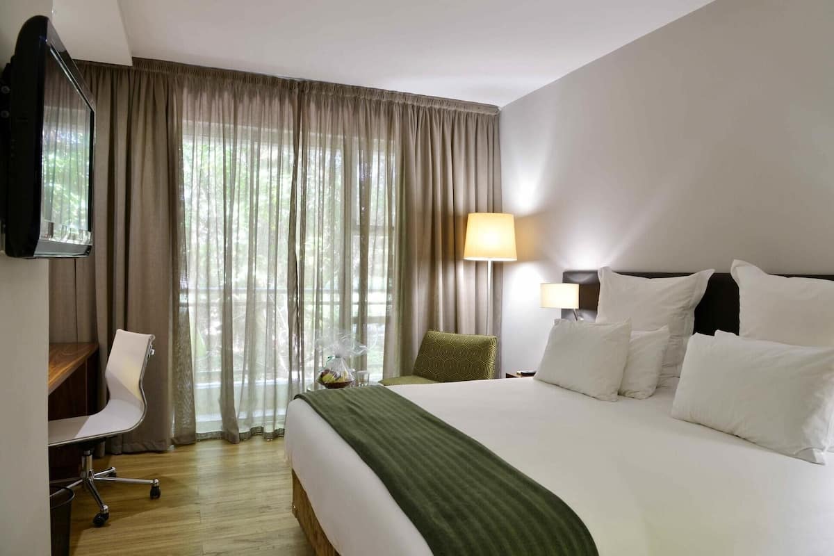 A hotel room with a large bed, a desk with a chair, a lamp, and a view of greenery through the window.