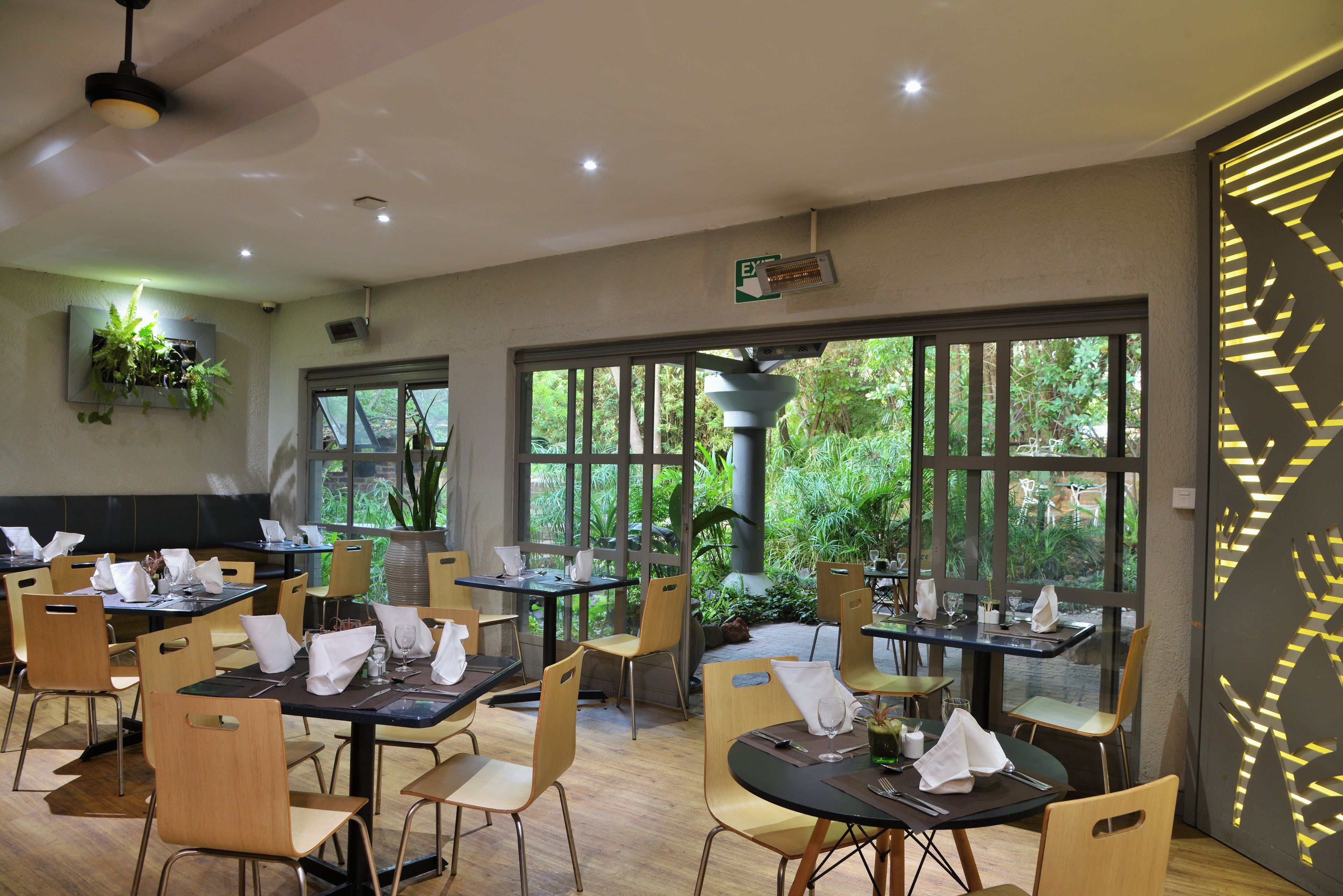 Un restaurante con mesas dispuestas para comer, ventanas amplias y plantas de interior.