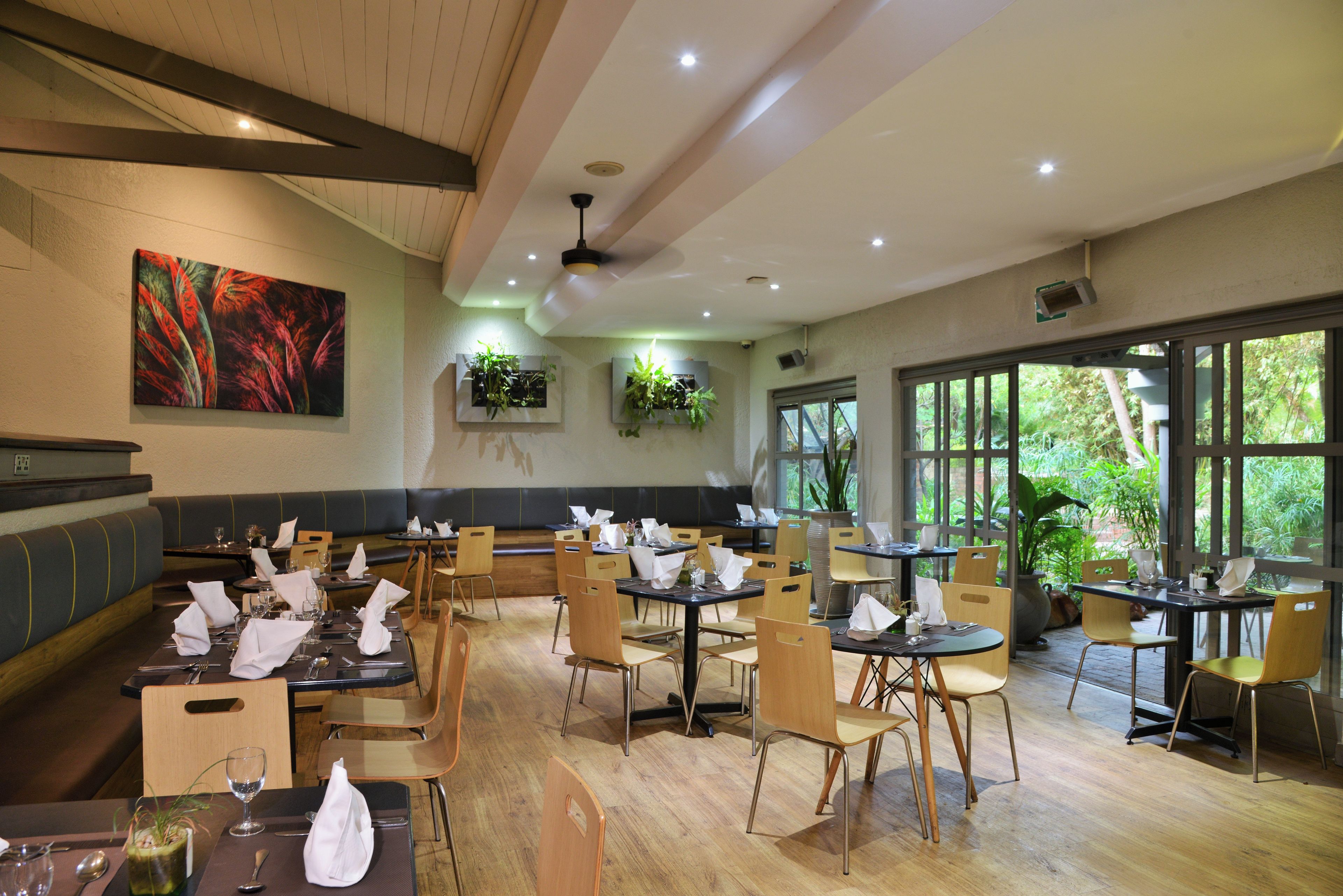 Un restaurante con varias mesas y sillas, un ventilador de techo y un ventanal con vista a la vegetación.