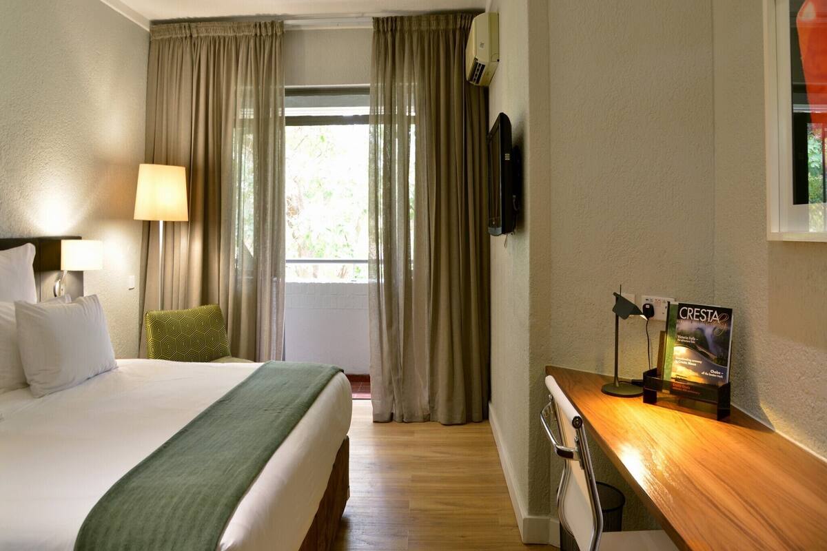 A hotel room with a bed, a desk, a lamp, a TV, and a window with curtains.