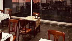 Een eetgedeelte met tafels en stoelen, grote ramen met uitzicht op de stad, en inbouwspots.