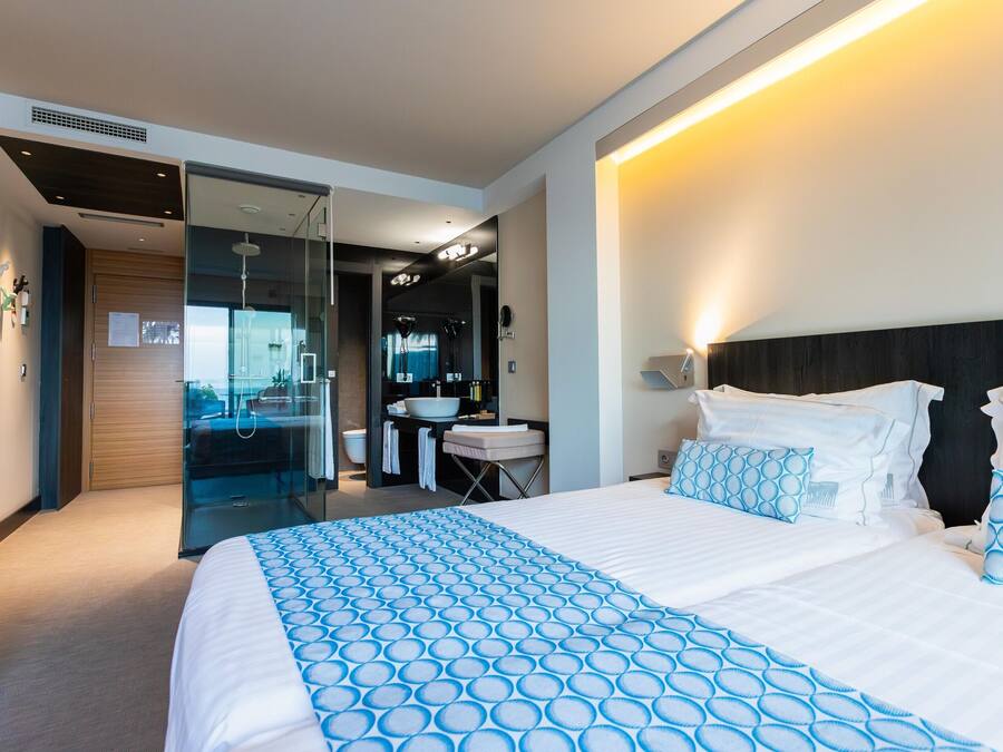 A modern hotel room with a large bed, a bathroom with a sink and mirror, and a view through a glass door.