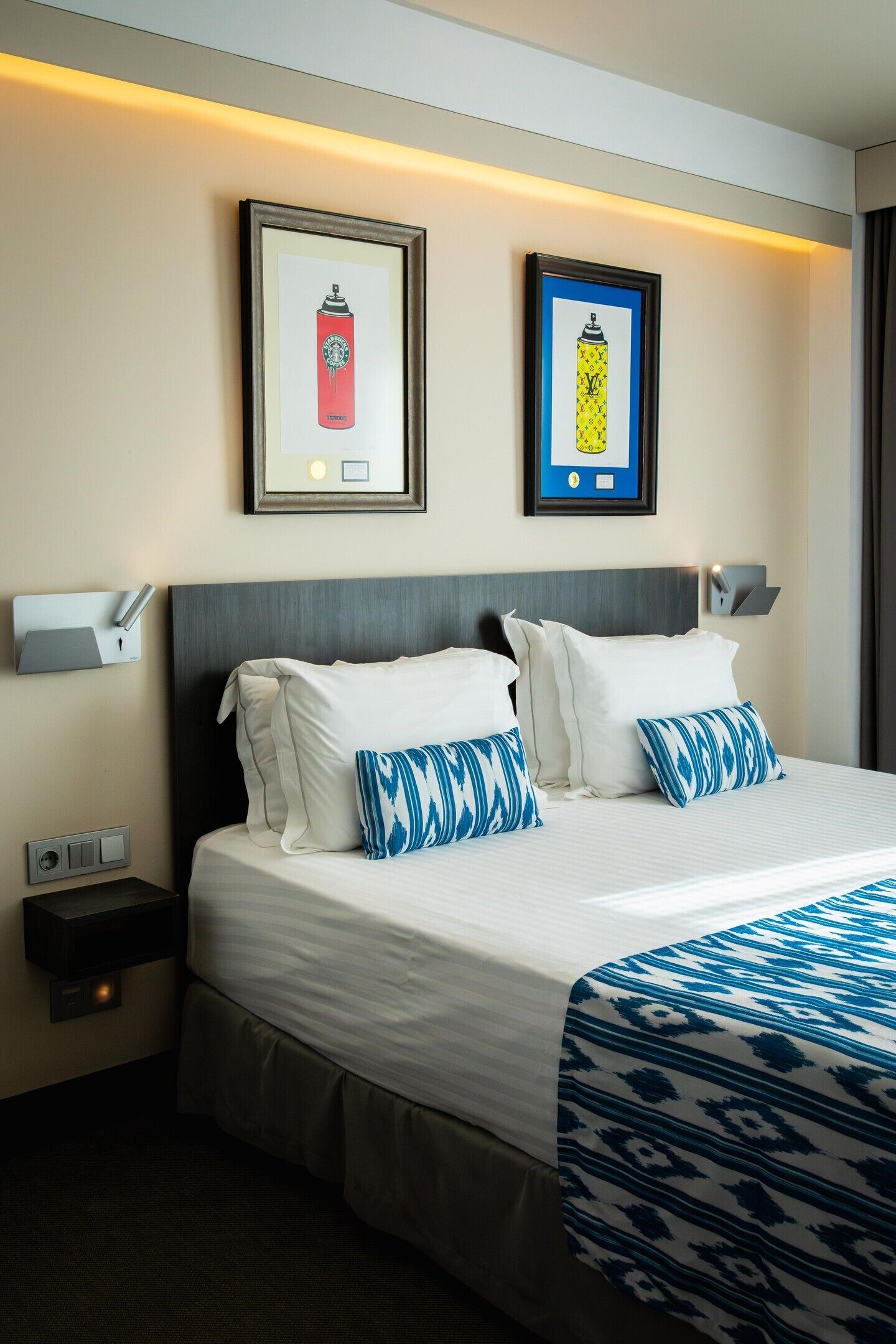 A hotel room with a bed, two pillows, a bedside table, a wall-mounted light, and two framed pictures.