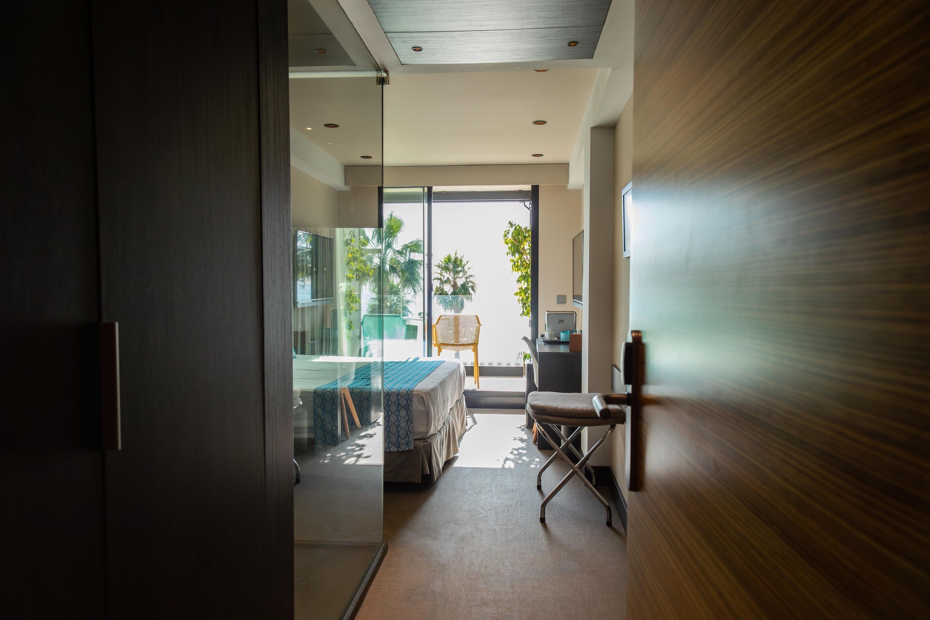 A modern hotel room with a glass door leading to a balcony, a bed, a desk, and a chair.