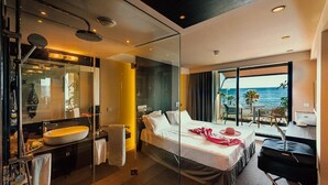 Una habitación de hotel moderna con una cama grande, cabina de ducha con paredes de vidrio, vista al mar y un balcón con mesa y sillas.