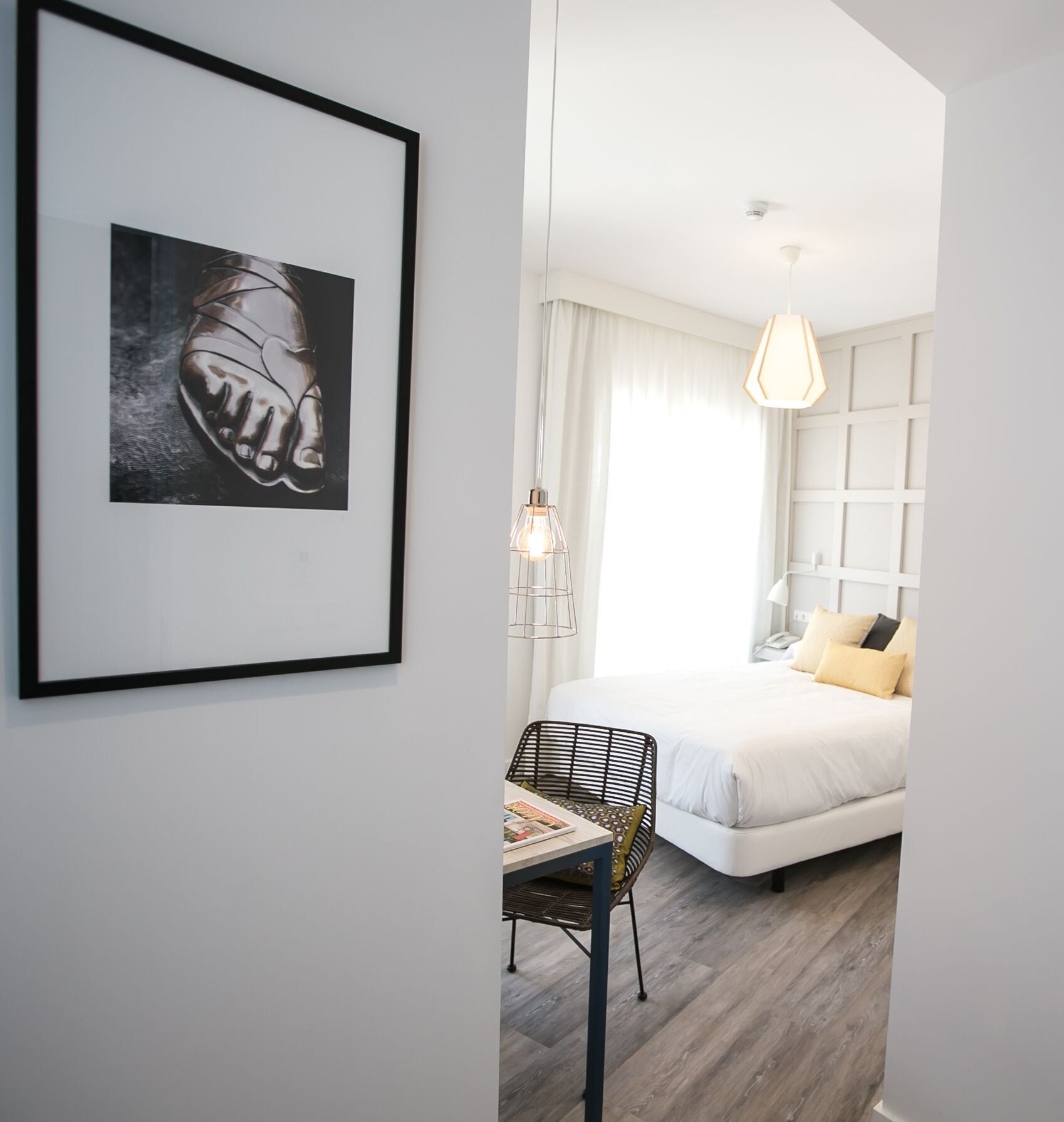 A modern hotel room with a large bed, a desk with a chair, a framed artwork on the wall, and a hanging light fixture.
