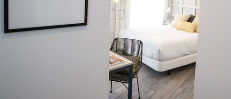A modern hotel room with a large bed, a desk with a chair, a framed artwork on the wall, and a hanging light fixture.