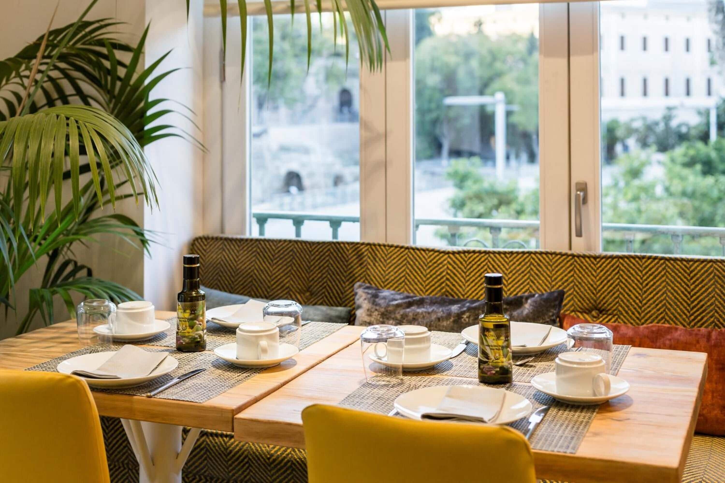 Comedor con mesa dispuesta para cuatro, un banco con cojines y vistas a zonas verdes y edificios.