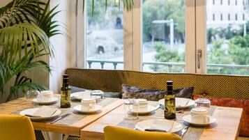 Comedor con mesa dispuesta para cuatro, un banco con cojines y vistas a zonas verdes y edificios.