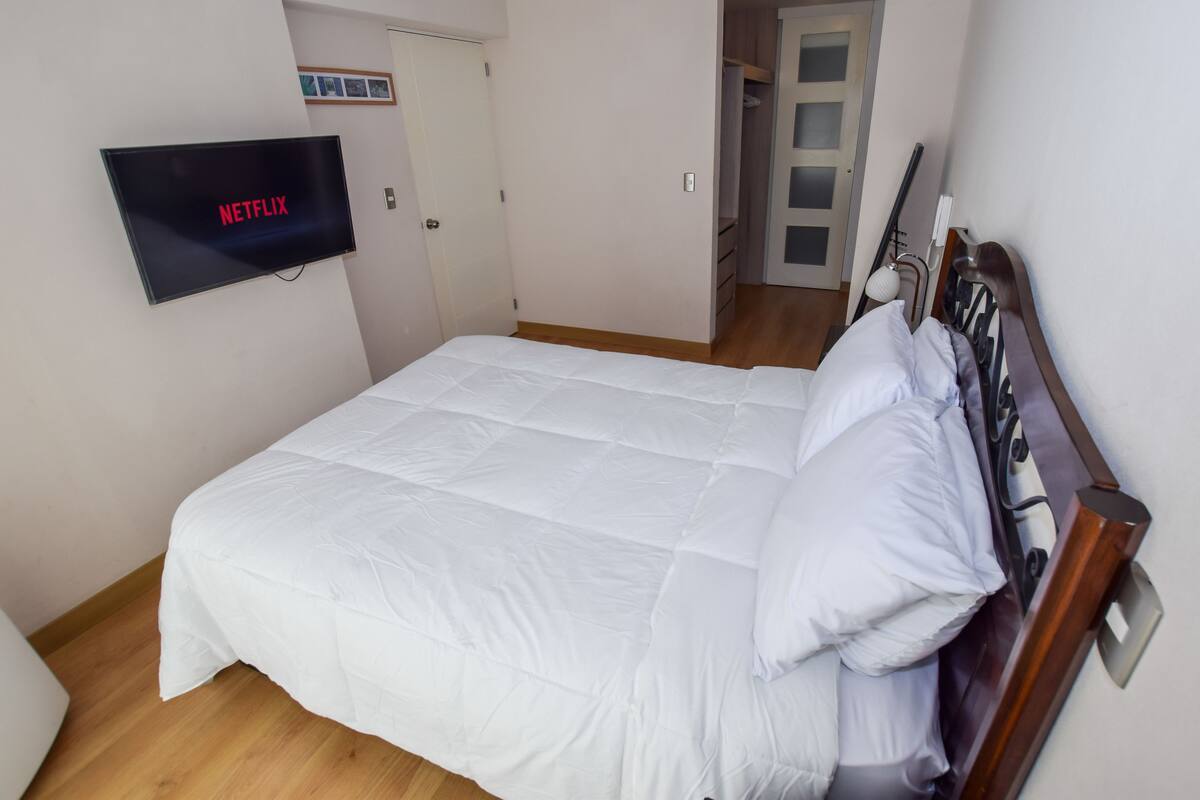 A neatly made bed with white bedding, a wall-mounted TV displaying the Netflix logo, and a wooden floor.