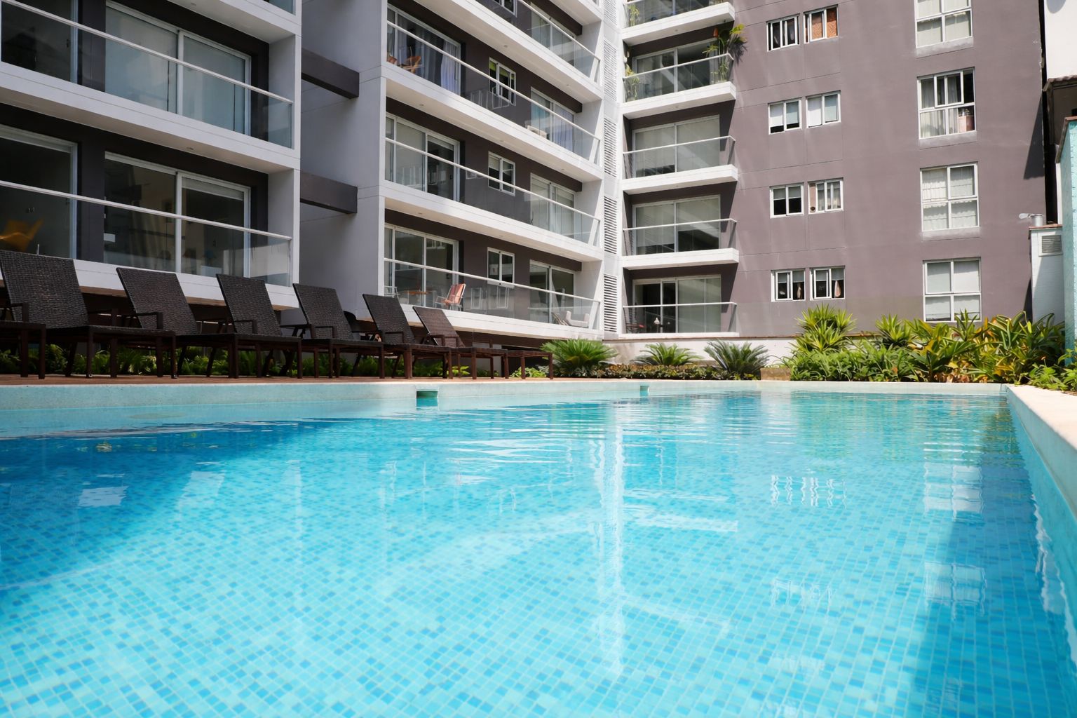 A swimming pool with lounge chairs, a multi-story building with balconies, and a clear blue sky.