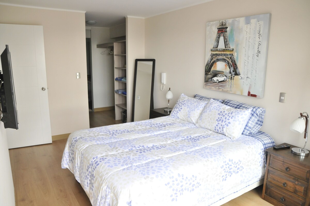 A bedroom with a bed, a nightstand, a mirror, a wardrobe, and a painting of the Eiffel Tower.