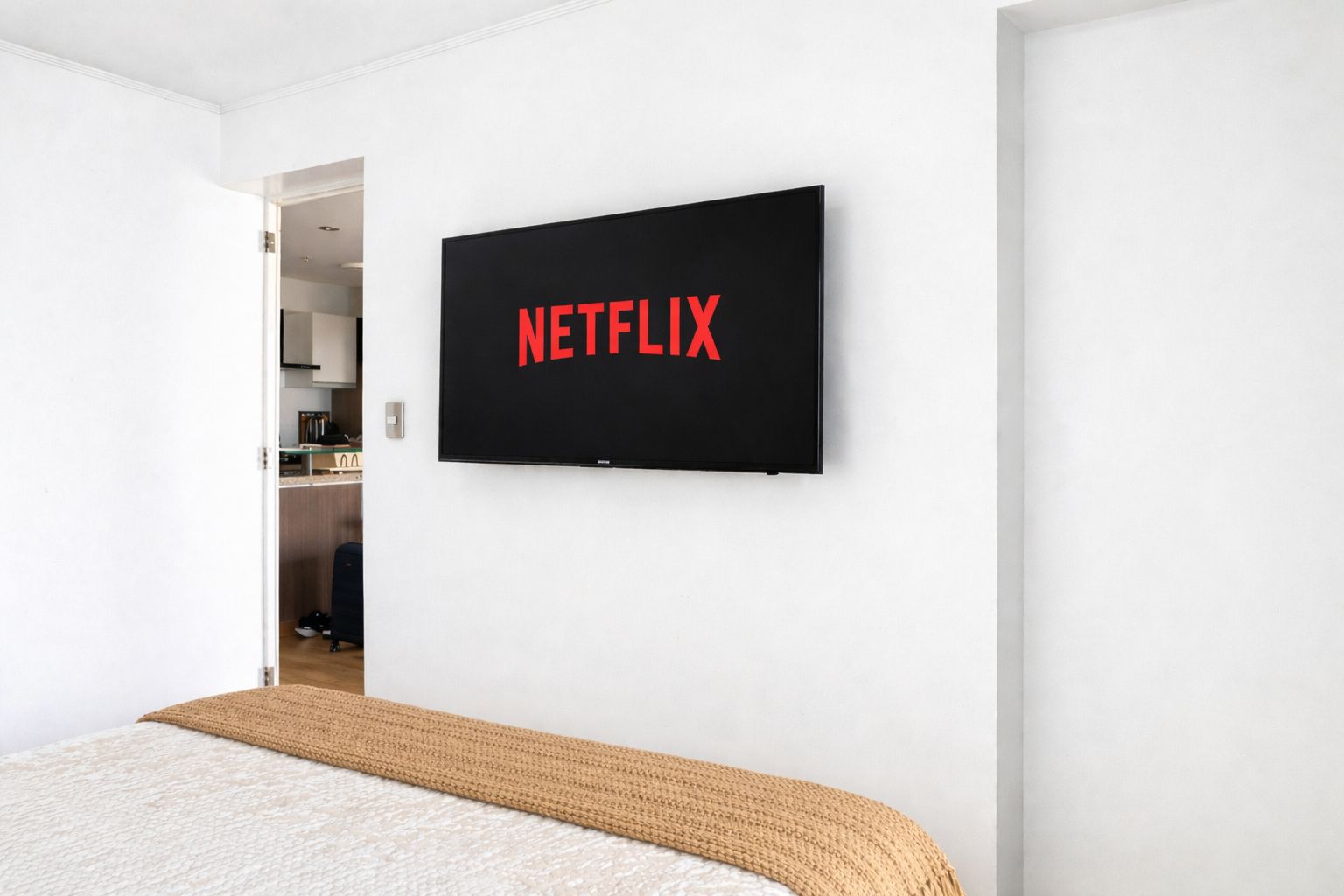 A modern hotel room with a bed, a flat-screen TV mounted on the wall displaying 'NETFLIX,' and a doorway leading to a kitchenette.