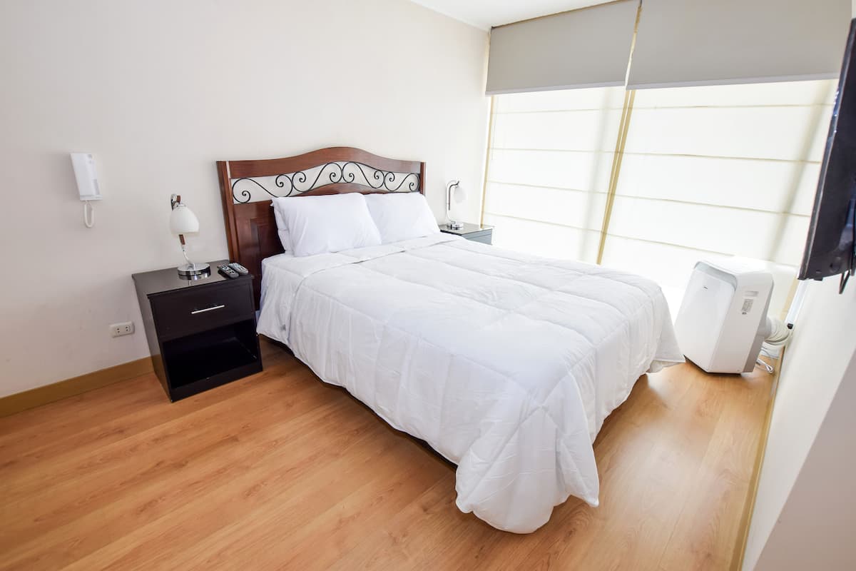 A neatly arranged bedroom with a wooden floor, a bed with white bedding, a bedside table with a lamp, a phone, and a heater.