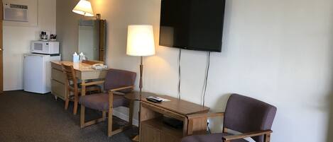 A hotel room with a desk, two chairs, a television, a lamp, a microwave, and a refrigerator.