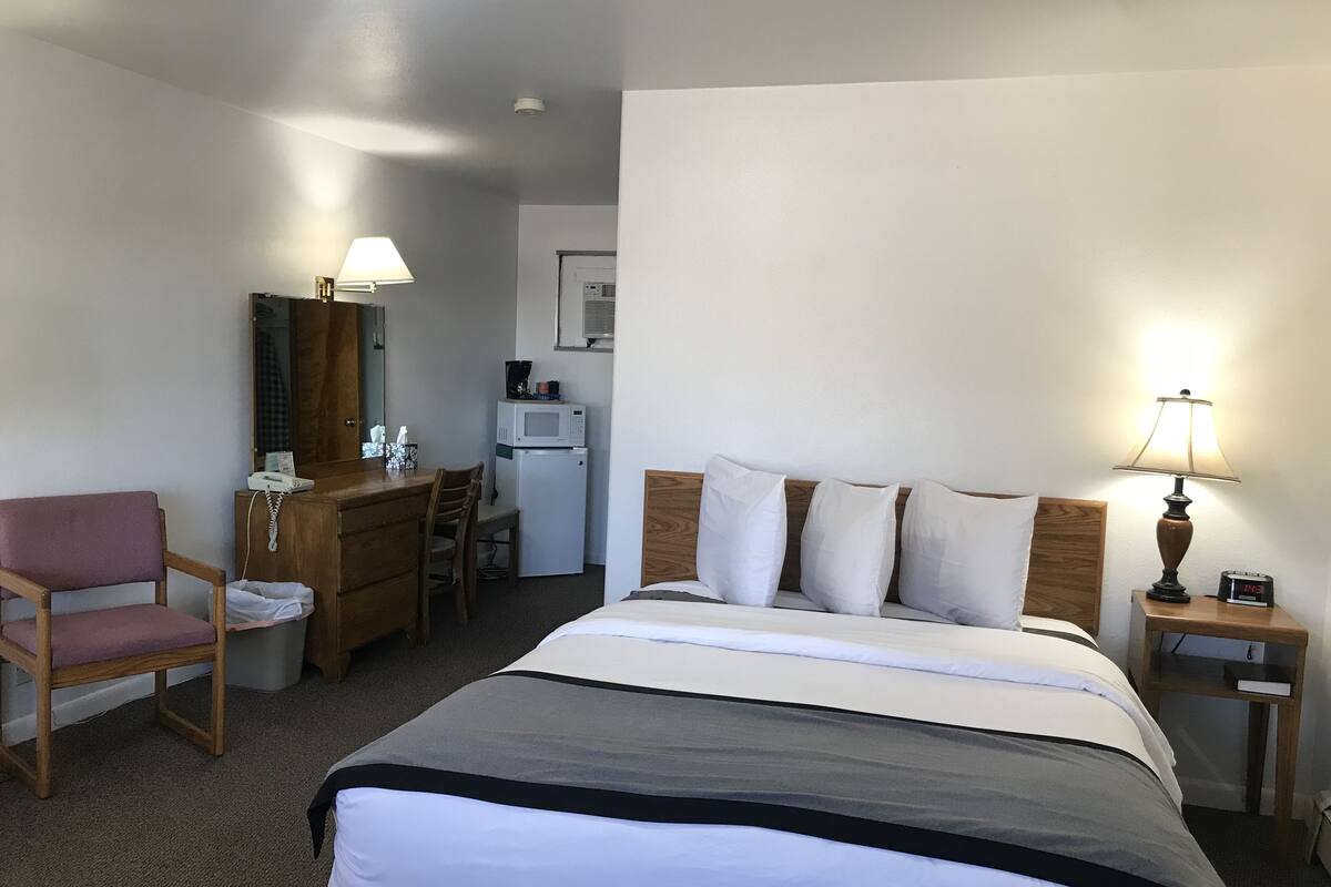 A hotel room with a bed, a desk, a chair, a lamp, and a clock.