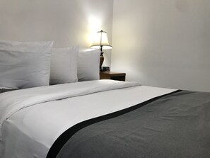 A neatly made bed with white bedding and a black accent, a bedside table with a lamp, and a plain wall. - Sage Motel (Newcastle)