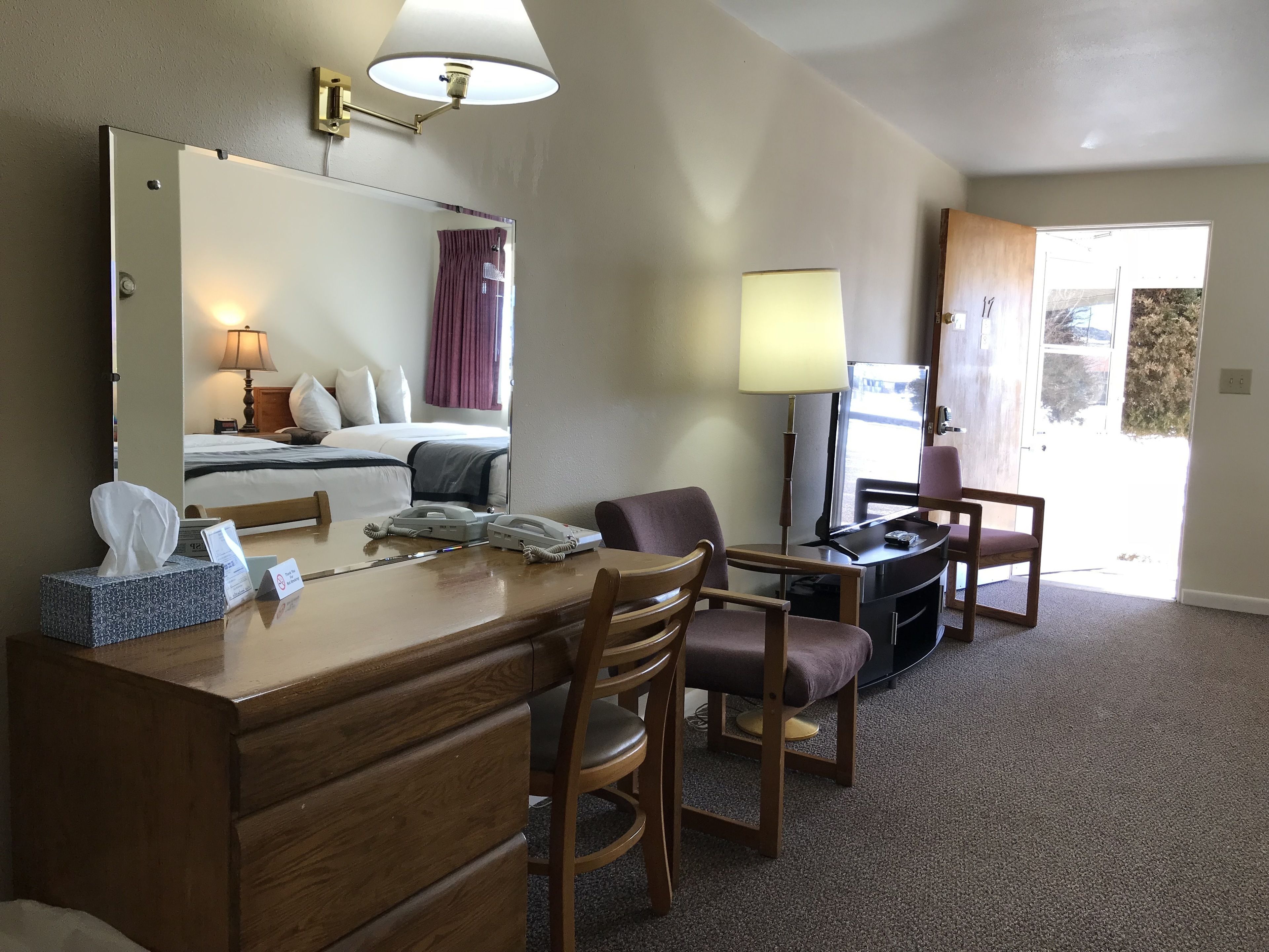 A hotel room with a wooden desk, two chairs, a bed with a headboard, a large mirror, a TV, and a door leading to a balcony.