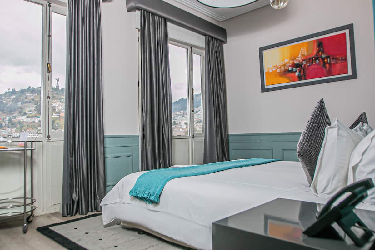 Quarto de hotel com cama, escrivaninha, cadeira, janela com cortinas, vista para a cidade e um quadro na parede.
