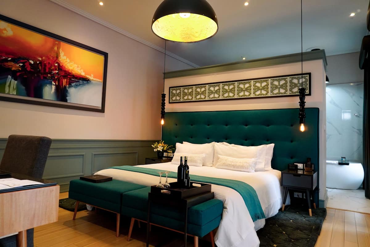 Quarto de hotel moderno com uma cama grande, uma escrivaninha, uma cadeira, um quadro e uma mesa de cabeceira.