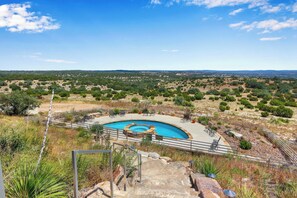 Una piscina de diseño circular, rodeada por un suelo de piedra y barandillas de metal, ubicada en un paisaje seco y montañoso con vegetación escasa.